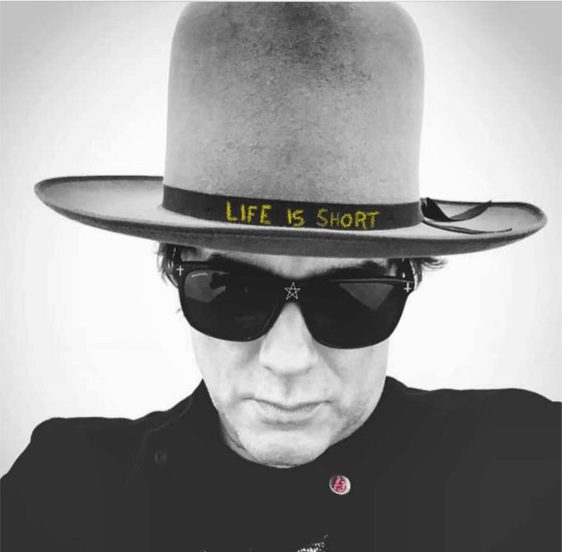 Black and white photo of Yul Vazquez in a hat that says "Life is short."