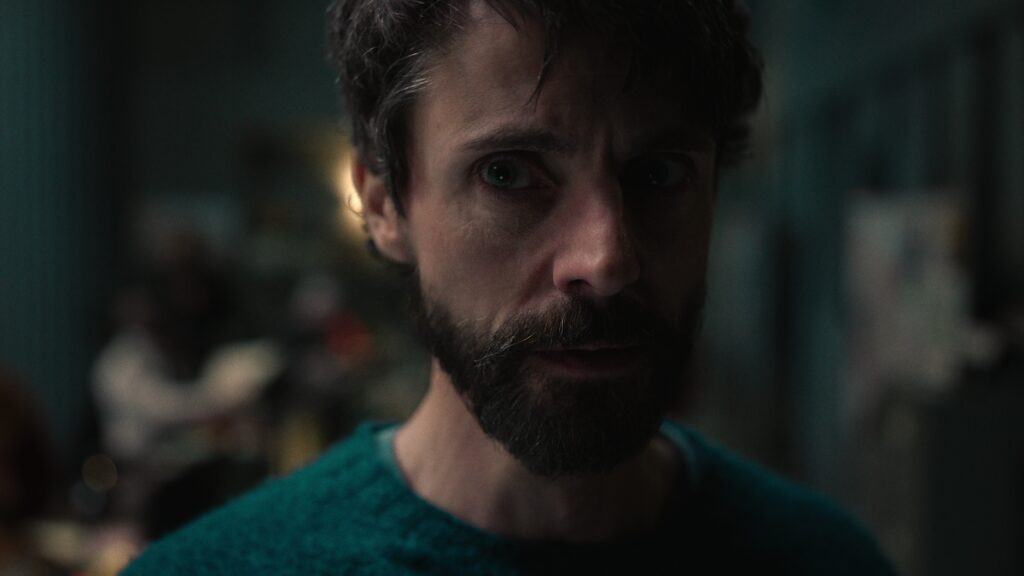Matthew Goode in a blue/green sweater looking concerned in an out of focus apartment.