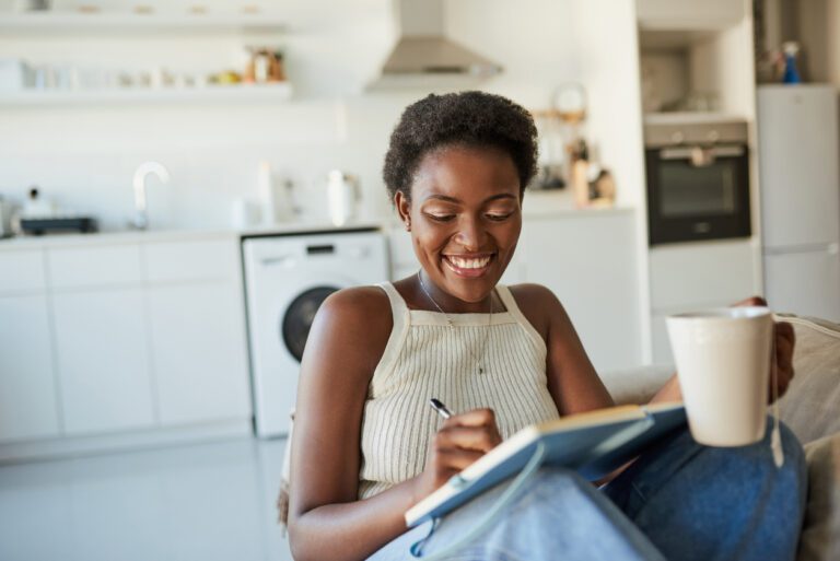 Young Black woman having tea in her home kitchen while happily jotting down notes in her notepad.